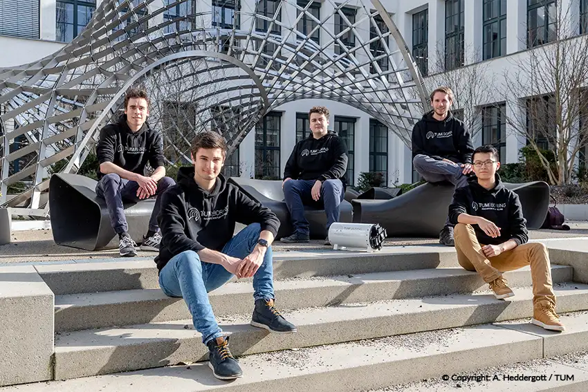 Fünf TUM Boring Mitglieder in passenden Kapuzenpullovern sitzen und posieren auf Außentreppen vor einem modernen Gebäude mit einer geometrischen Metallskulptur darüber.