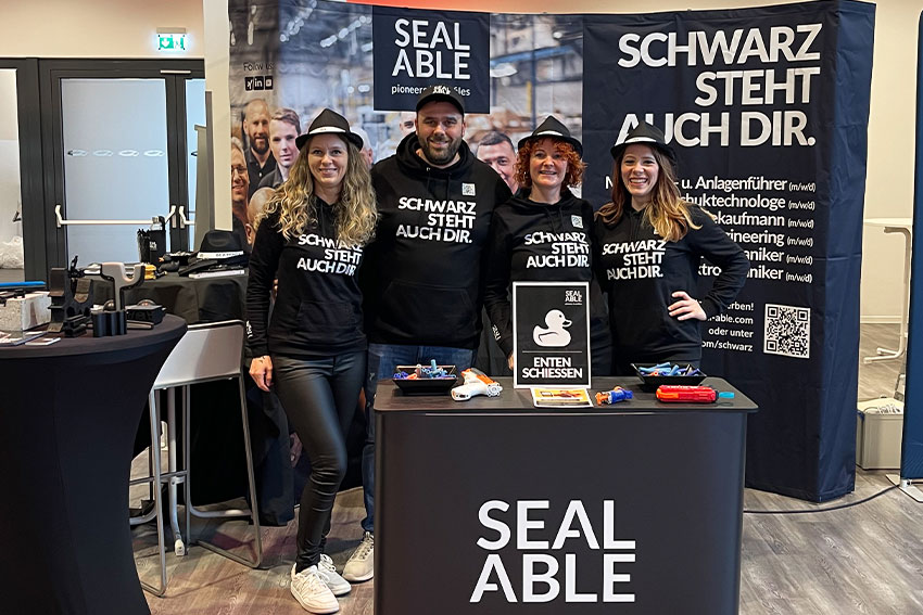 Vier Mitarbeiter stehen am Stand zur Jobmesse und verkörpern das Motto "Schwarz steht auch dir"